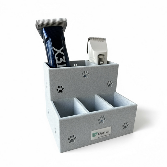 A gray Clip Sharp Large Storage Caddy with paw print cutouts holds two electric pet clippers, features several empty compartments, and sits against a plain white background. The front label reads “ClipSharp.”.
