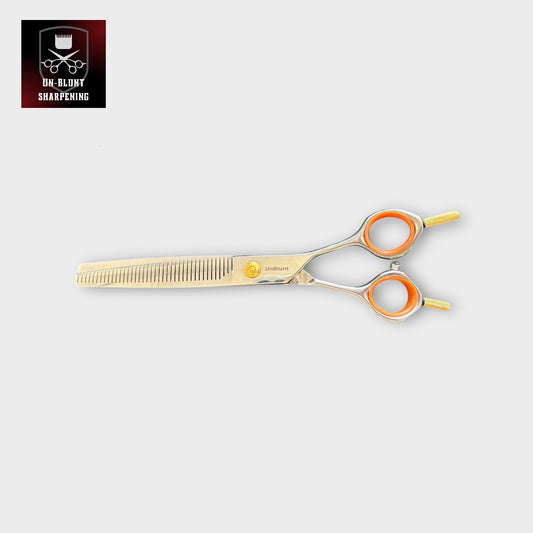 The UnBlunt Straight Piano Teeth Fluffers 7" Scissors, featuring silver and gold tones, orange finger rings, and fine-tooth thinner blades, are displayed on a light gray background with the UN-BLUNT SHARPENING logo in the top left corner.