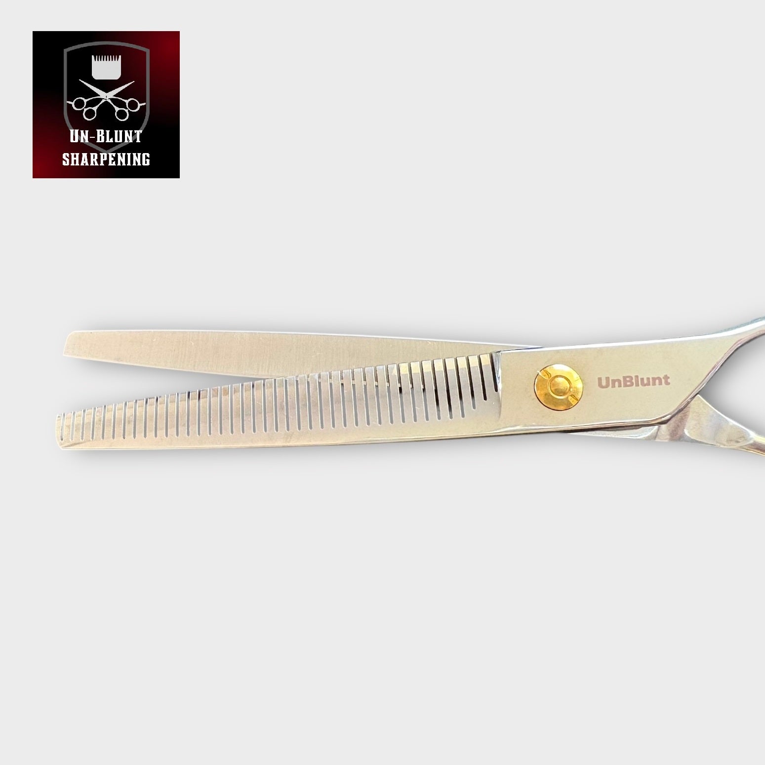 UnBlunt Straight Piano Teeth Fluffers 7" Scissors—silver thinning shears with a gold screw on a light backdrop. A black and red UN-BLUNT SHARPENING logo with comb and scissors icon appears. Ideal for precise, fine-tooth hair styling.