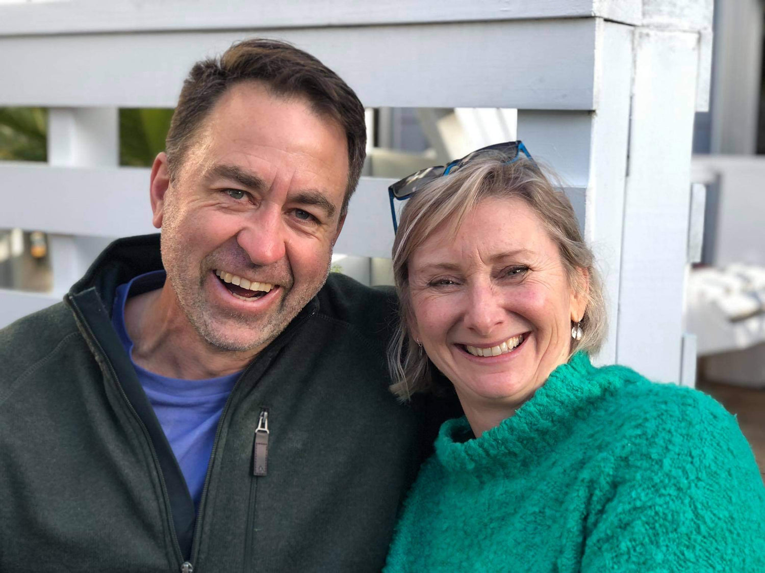 A smiling man in a dark jacket sits next to a smiling woman in a green sweater. They are outdoors, with a white fence and sunlight in the background. Both look happy and relaxed.